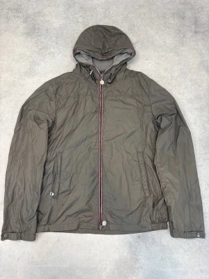 Grå vindjacka med huva från Moncler - Snygg grå vindjacka från Moncler med huva och hel dragkedja framtill. Jackan har två sidofickor med dragkedja och ribbad grå insida vid kragen. Detaljer med Monclers signaturrand längs dragkedjan och huvkanten. Perfekt för blåsiga dagar.