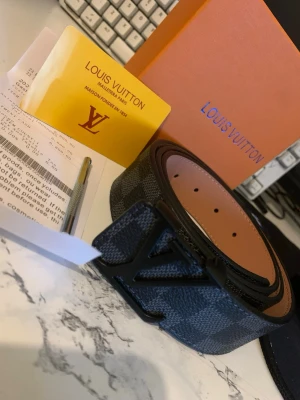 L,V bälte i skinn - Snyggt bälte från L,V i svart och grått med klassiskt rutigt mönster och stor L,V-logga i svart metall. Tillverkat i exklusivt skinn och levereras med originalbox och certifikat. Perfekt accessoar för att lyfta din outfit.