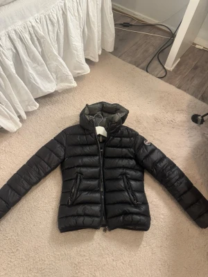 Svart dunjacka med huva från Colmar - Snygg svart dunjacka från Colmar med huva och två dragkedjeförsedda fickor framtill. Jackan har quiltad design och en diskret logga på ärmen. Perfekt för kalla vinterdagar och riktigt lätt att bära. Materialet är glansigt och ger en modern look. Och har två skador som finns på bilderna 