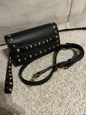 Svart med nitar - Snygg svart i läderimitation med guldfärgade nitar och detaljer. Väskan har både handledsrem och avtagbar axelrem, vilket gör den flexibel att bära. Rektangulär form och aldrig använd !