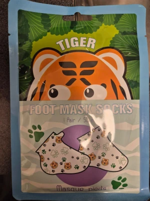 Tiger Foot Mask Socks - Ett par fotmask-strumpor från Tiger, designade med söta tigeransikten och gröna tassavtryck. Förpackningen är färgglad med ett tigeransikte och gröna blad. Perfekt för att återfukta och vårda fötterna på ett roligt sätt.