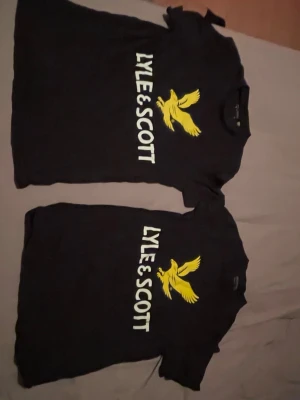 (BUNDLE). Svart Lyle & Scott t-shirt med logga - (BUNDLE). Säljer en svart t-shirt från Lyle & Scott med stor vit logotyp och gul fågel på bröstet. Klassisk rund hals och korta ärmar. Perfekt för dig som gillar streetwear och vill ha en enkel men snygg look.