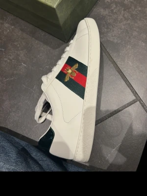 Gucci Ace vita sneakers med bi - Stilrena vita sneakers från Gucci med klassiska gröna och röda ränder på sidan samt en broderad guldfärgad bi. Skon har vit sula med unikt mönster undertill och detaljer i mörkgrönt vid hälen. Tillverkad i läder.