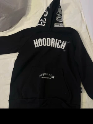 Svart Hoodrich set med tryck - Säljer ett svart Hoodrich set med matchande hoodie och byxor. Hoodien har stor vit logga på bröstet och tryck på huvan, samt känguruficka. Byxorna har vit Hoodrich-tryck över bakdelen och liten logga på benet. Mjukt material, perfekt för chill eller streetwear.