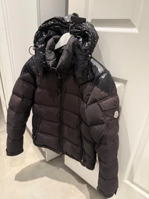 Moncler chevalier down jacket  - Snygg svart dunjacka från Moncler med glansiga detaljer på axlar och huva. Jackan har en hög krage, avtagbar huva och två dragkedjefickor framtill. Klassisk Moncler-logga på ärmen och quiltad design för extra värme. Finns lite små defekter så som carton borta jackan är äkta och bevis finns jackan är lite sol blekt samt använd vilket du ser på bild annars väldigt bra sick 