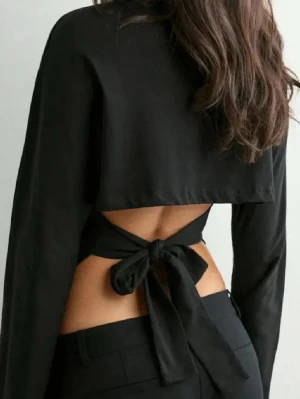 Svart topp med öppen rygg och knyt - Trendig svart topp med långa vida ärmar och öppen rygg som knyts med en stor rosett. Toppens material är mjukt och stretchigt, vilket ger en snygg passform. Perfekt för dig som vill ha en unik och stilren look.