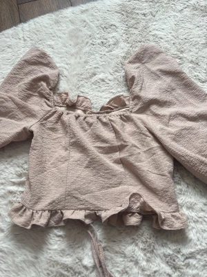 Beige blus med volang och snörning - Supersöt beige blus med puffiga långärmar, volangkant nedtill och snörning framtill. Tillverkad i ett strukturerat, lätt material som ger en romantisk vibe. Perfekt för dig som gillar trendiga detaljer och en feminin look.