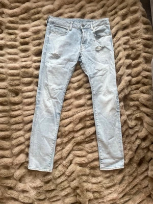 Ljusblå slitna skinny jeans - Säljer ett par ljusblå skinny jeans med slitningar vid knäna och klassisk femficksdesign. Jeansen har en smal passform och är tillverkade i denim med lite stretch för extra komfort. Perfekta för en avslappnad och trendig look.storleken ör w32/l32