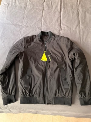 Ny Svart bomberjacka från Style - Snygg svart bomberjacka i storlek M. Jackan har ribbade muddar vid ärmslut och midja, två snedställda fickor med knappdetaljer och dragkedja framtill. Klassisk bombermodell med stilren look, perfekt till streetwear eller casual outfits. Helt ny aldrig använd 