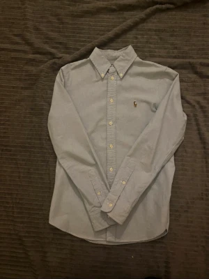 Ljusblå skjorta från Ralph Lauren - Klassisk ljusblå skjorta från Ralph Lauren med button-down krage och broderad logga på bröstet. Skjortan har långa ärmar och vita knappar framtill. Perfekt för en stilren och tidlös look.
