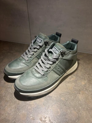 Gröna sneakers från BORG - Stilrena gröna sneakers från BORG med grå snörning och vit sula. Skorna har en modern design med logga på sidan och plös, samt detaljer i syntetläder. Perfekta för dig som vill ha en clean look till vardagsoutfiten.