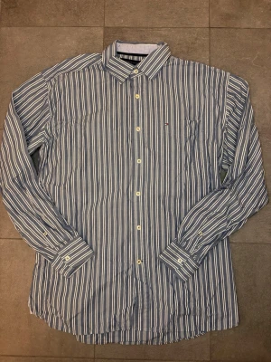 Randig skjorta från Tommy Hilfiger - Snygg blå och vit randig skjorta från Tommy Hilfiger med klassisk krage och bröstficka. Skjortan har långa ärmar, knappar framtill och diskret logga på fickan. Perfekt för dig som gillar stilrena och tidlösa plagg.