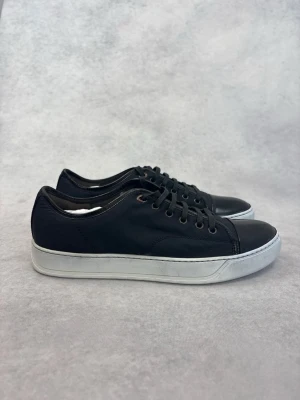 Svarta låga Lanvin sneakers med vit sula - Stilrena svarta sneakers med vit sula och svarta skosnören. Ovandelen är i textil och tån är i blankt material som ger en snygg kontrast. Perfekta för en clean och modern look.
