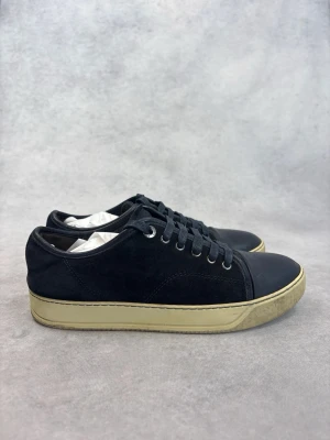 Svarta låga Lanvin sneakers med beige sula - Stilrena svarta sneakers med lågt skaft och beige sula. Ovandelen är i mocka och tåpartiet i slätt läder. Klassisk snörning med svarta skosnören och metallöljetter. Perfekta för en clean och avslappnad look.