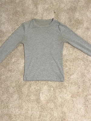 Grå långärmad topp i bomull - En enkel och stilren grå långärmad topp i mjuk bomull. Modellen är figurnära med rund halsringning och passar perfekt till jeans eller kjol. Perfekt basplagg för din garderob.