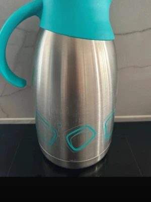 Termoskanna i rostfritt stål turkos - Säljer en termoskanna i rostfritt stål med turkost plastlock och handtag. Kannan har en rundad form och dekoreras med turkosa geometriska mönster på sidan. Perfekt för att hålla drycker varma eller kalla under längre tid.