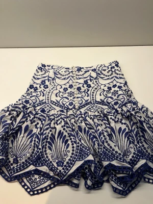 Vit och blå broderad kortkjol - Supersnygg vit kortkjol med blått broderat mönster och dekorativa knappar framtill. Kjolen har volangkant och ett unikt, detaljerat blommönster som verkligen sticker ut. Perfekt för dig som vill ha något extra till sommargarderoben!