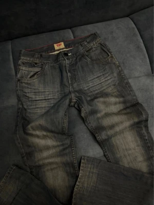 Grå slitna Levi's jeans - Säljer ett par grå Levi's jeans med snyggt sliten tvätt och klassiska bakfickor med V-formad söm. Jeansen har fem fickor, bälteshällor och knappgylf. Tillverkade i bomull med en cool vintagekänsla. Perfekta för en avslappnad streetstyle. Bredd 70 längd 102