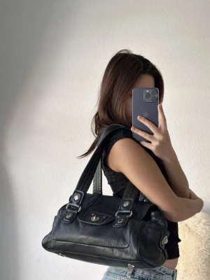 Svart axelremsväska i skinn - Snygg svart handväska i mjukt skinn med dubbla axelremmar och silverfärgade detaljer. Väskan har ett lock med vridlås framtill och dekorativa sömmar längs sidorna. Perfekt storlek för det viktigaste och ger en cool vibe till din outfit.