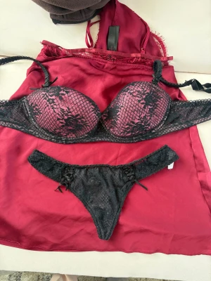 Svart spets-bh och trosa set la vie en rose  - Säljer ett snyggt underklädesset med svart spets-bh och matchande trosa. Bh:n har vadderade kupor med spetsdetaljer och justerbara axelband. Trosan är i svart spets med små rosetter på sidorna. Båda delar har en lyxig känsla och ligger på ett vinrött nattlinne i satin.