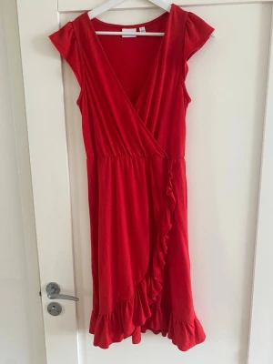 Röd omlottklänning från Vila - Snygg röd omlottklänning från Vila med volangdetaljer längs nederkanten och omlottskärning framtill. Klänningen har korta ärmar och markerad midja för en feminin siluett. Perfekt för sommarens alla tillfällen.