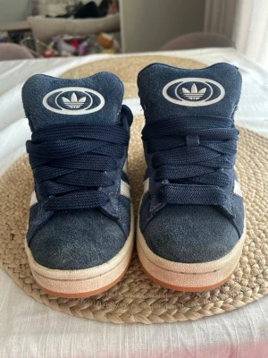Adidas Campus mörkblå sneakers - Mörkblå Adidas Campus sneakers i mocka med klassiska vita ränder på sidan och Adidas-logga på plösen. Skorna har en låg profil och vit sula med gummidetaljer. Snygg retrostil som passar perfekt till streetwear och vardagsoutfits. Storlek 36 och en halv. 