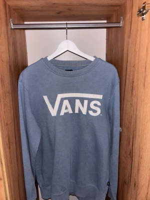 Blå sweatshirt från Vans - Säljer en blå sweatshirt från Vans med stort vitt logotryck på bröstet. Tröjan har rund halsringning, långa ärmar och ribbade muddar vid ärmslut och nederkant. Perfekt för en avslappnad streetwear-look.