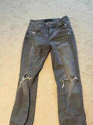Grå slitna jeans med raka ben - Säljer ett par grå jeans med raka ben och flera slitningar, bland annat vid knäna och fickorna. Jeansen har klassisk femficksdesign och är gjorda i denim. Perfekta för en avslappnad och trendig look. (Hålen är med flit, köpta så)