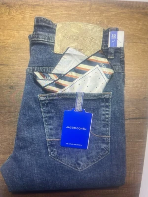 Blå jeans från Jacob Cohën - Snygga blå jeans från Jacob Cohën med klassisk femficksdesign och ljus tvätt. Jeansen har en beige patch med logga bak och levereras med en färgglad scarf i fickan. Perfekt passform och exklusiv känsla.