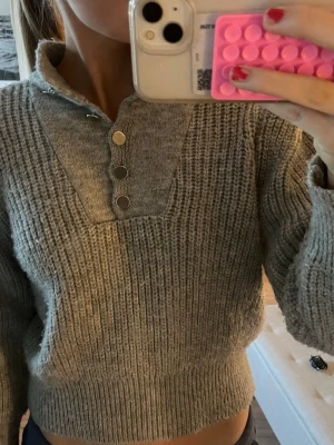 Grå stickad tröja med knappar - Mysig grå stickad tröja med ribbad struktur och hög krage. Tröjan har tre silverfärgade tryckknappar framtill och breda muddar vid ärmslut och nederkant. Perfekt för kalla dagar och enkel att matcha med olika outfits.