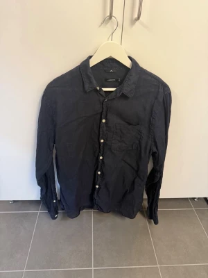 Mörkblå linneskjorta från J.Lindeberg, stl M - Snygg mörkblå skjorta från J.Lindeberg i luftigt linne. Klassisk krage, bröstficka och vita knappar framtill. Perfekt till både jeans och chinos för en clean och stilren look.