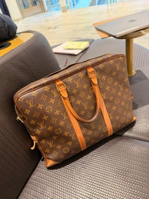 Louis Vuitton Monogram datorväska - Snygg datorväska från Louis Vuitton i klassiskt brunt monogrammönster med ljusbruna läderhandtag och detaljer. Väskan har dragkedja, flera innerfack och är rymlig nog för laptop och tillbehör. Perfekt för dig som vill ha en lyxig och stilren look. 