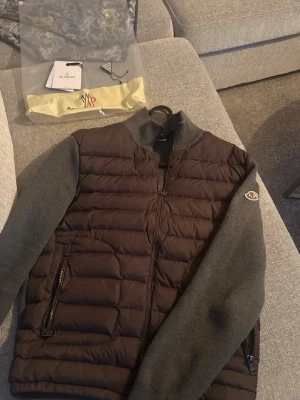 Moncler cardigan grå - Snygg cardiganjacka från Moncler med vadderad brun front och grå stickade ärmar. Jackan har dragkedja framtill, två sidofickor med dragkedja och klassisk Moncler-logga på ärmen. Perfekt för lager-på-lager och streetwear-stil. Storlek 2 (M)