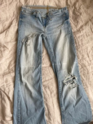 Ljusblå slitna bootcut jeans - Säljer ett par ljusblå bootcut jeans från Nelly med egengjorda slitningar och hål på både lår och knä. Jeansen har rå kant upptill, klassiska fem fickor och är tillverkade i denim. Perfekta för en avslappnad och trendig look. Skriv om ni har andra frågor!🥰