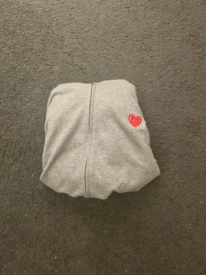Grå hoodie från Comme des Garçons Play - Säljer en grå hoodie från Comme des Garçons Play med dragkedja och det ikoniska röda hjärtat med ögon på bröstet. Tillverkad i mjukt bomullsmaterial och har en klassisk, avslappnad passform. Perfekt för dig som gillar streetwear och stilrena detaljer.