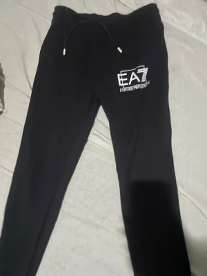 Svarta EA7 mjukisbyxor - Svarta mjukisbyxor från Emporio Armani EA7 med dragsko i midjan och vit logga på vänster lår. Sköna och stilrena med smal passform, perfekta för chill eller träning. Tillverkade i mjukt material för maximal komfort. Även köpt från johnells för cirka 1299kr