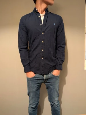 Mörkblå skjorta från Ralph Lauren - Klassisk mörkblå skjorta från Ralph Lauren med button-down krage och broderad logga på bröstet. Skjortan har långa ärmar, vita knappar och är tillverkad i bomull. Perfekt till jeans eller chinos för en clean look.