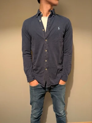 Mörkblå skjorta från Ralph Lauren - Klassisk mörkblå skjorta från Ralph Lauren med broderad logga på bröstet. Skjortan har långa ärmar, knappar framtill och är tillverkad i mjuk bomull. Perfekt för en stilren och avslappnad look.