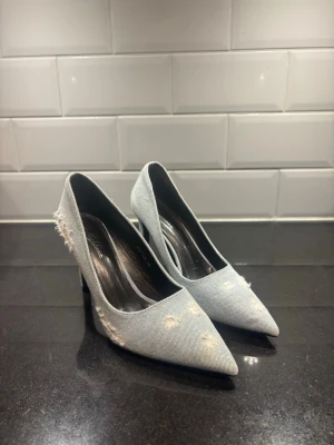Ljusa denim pumps med slitningar - Säljer ett par ljusblå pumps i denimtyg med coola slitningar och fransade detaljer. Skorna har spetsig tå och 9cm höga klackar som ger en edgy look. Perfekt för dig som gillar trendiga och unika skor.