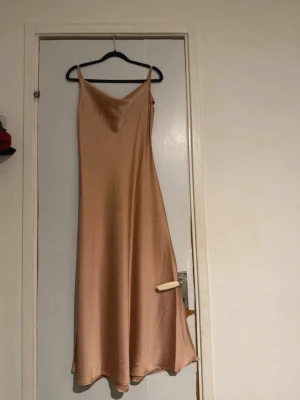 Champagnefärgad långklänning i satin - Säljer en elegant långklänning i champagnefärgad satin med smala axelband och v-ringad halsringning. Klänningen har en mjuk, glansig yta och ett stilrent, minimalistiskt snitt som ger en lyxig känsla.