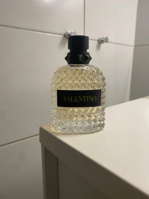 Valentino parfym uomo born in Roma yellow edt 100ml - Snygg parfym från Valentino i en genomskinlig glasflaska med nitar och rutmönster. Flaskan har en svart etikett med guldfärgad text och ett coolt svart lock med nitar. Perfekt för dig som gillar exklusiv design och vill ha en parfym som sticker ut i badrumshyllan.