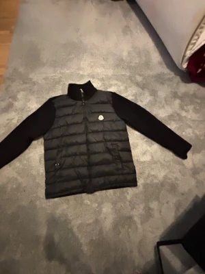 Svart dunväst från Moncler - Snygg svart dunväst från Moncler med quiltad front och ribbstickade ärmar. Västen har dragkedja, hög krage och Moncler-logga på bröstet. Perfekt för lager-på-lager och ger en clean look till din outfit.