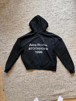 Svart hoodie Acne Studios Stockholm - Svart hoodie från Acne Studios med stor vit text på ryggen: 'Acne Studios STOCKHOLM 1996'. Klassisk passform med huva och ribbade muddar. Perfekt för dig som gillar streetwear och vill ha en stilren look med tydlig logga.