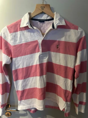 Rosa och vit randig rugbytröja Polo Ralph Lauren - Klassisk långärmad rugbytröja från Polo Ralph Lauren i breda rosa och vita ränder. Tröjan har krage, knappslå och den ikoniska broderade loggan på bröstet. Tillverkad i mjuk bomull för en bekväm och avslappnad stil.