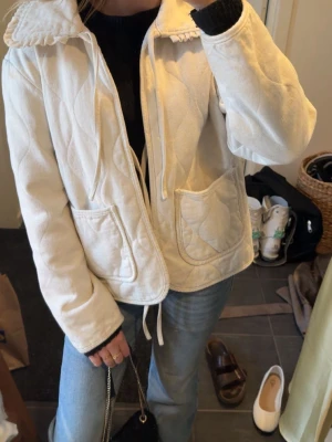 Vit quiltad jacka med volangkrage - Supersöt vit quiltad jacka med volangdetaljer på kragen och stora fickor framtill. Jackan har dragsko i midjan och är långärmad med en mjuk, mysig känsla. Perfekt för dig som vill ha en feminin och trendig look till vardags. Jackan har inte några slitningar men den har små sminkfläckar som lätt kan tvättas bort med tvål och vatten.