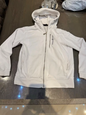 Vit hoodie från Sail Racing - Snygg vit hoodie från Sail Racing med dragkedja framtill och broderad logga på bröstet. Hoodien har två fickor på sidorna och en justerbar huva. Tillverkad i mjukt bomullsmaterial som känns skönt mot huden. Perfekt för en avslappnad och sportig stil. Lite slitage på båda armbågarna.