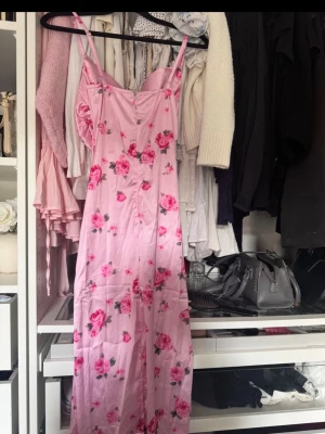 Lång rosa blommig klänning - Söt långklänning i ljusrosa med blommigt mönster. Klänningen har smala axelband och en figurnära passform med lätt glansigt material som ger en elegant känsla. Perfekt för sommarens alla tillfällen.