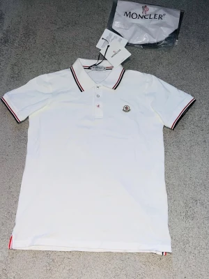 Vit Moncler Piké - Färg: Vitt 📝 Storlek: Medium(M)  📏 Mott: Axel till axel 48 Cm--Midja till hals 64 Cm  ♻️ Använd fåtal gånger-Nytt skick  📦 Postar inom 24h  Hör av er vid funderingar eller prisförslag!