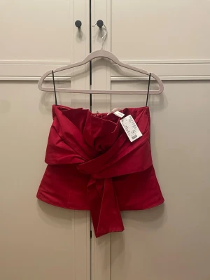 Röd axelbandslös topp med rosett - Snygg röd axelbandslös topp från H&M med stor dekorativ rosett framtill. Toppen har bandeau-modell och dragkedja i sidan för enkel på- och avtagning. Fin passform och kvalitet. Helt ny med prislapp. Storlek M men liten i stolek, passar en S. Dragkedja baktill