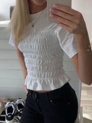 Vit smockad topp - Säljer en vit topp med smockad midja och volangkant nedtill. Toppen har korta ärmar och rund halsringning, vilket ger en söt och trendig look. Perfekt att styla med både jeans och kjol för en fräsch outfit.